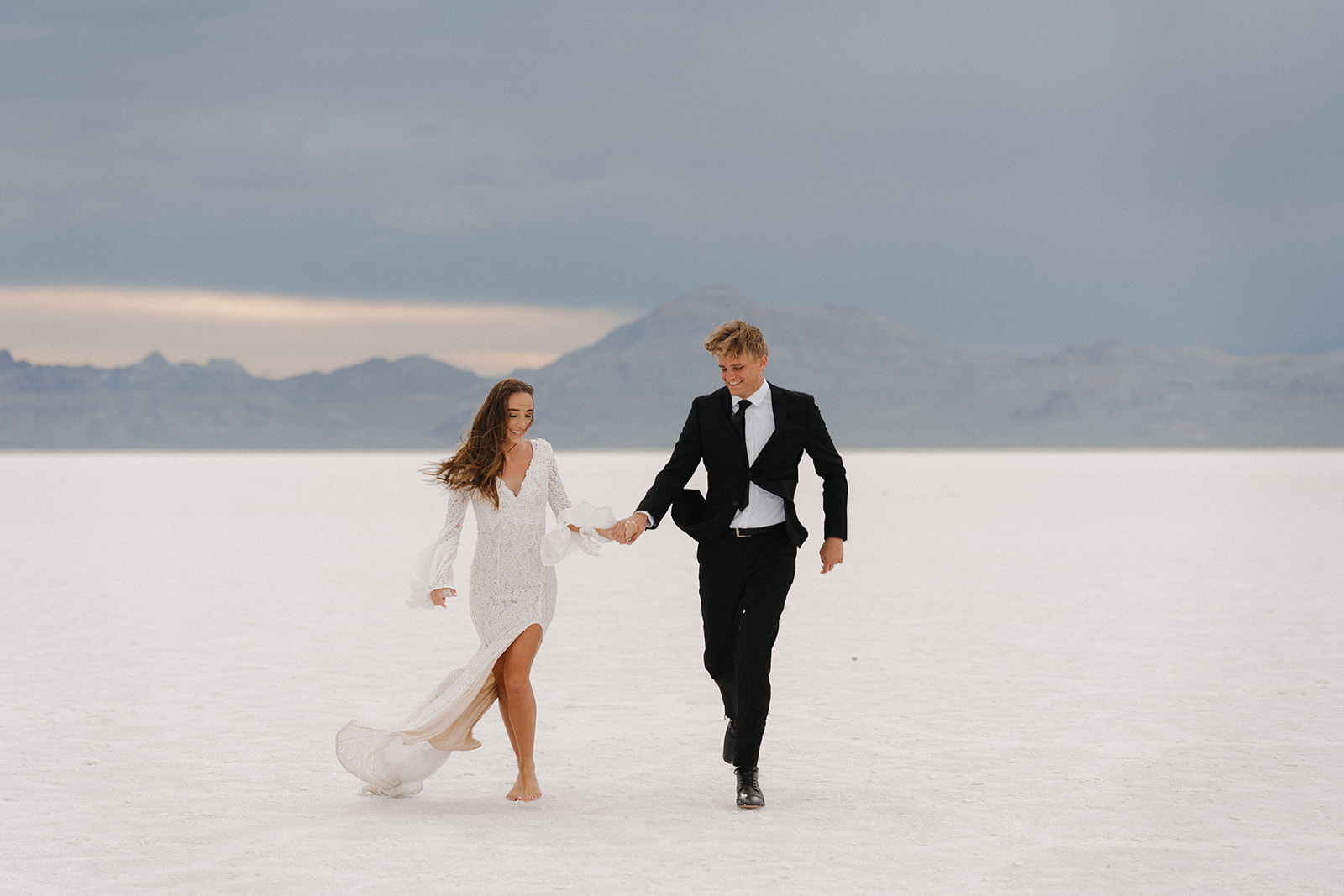 Newlyweds walk through the flats on a windy day in a black suit and lace dress during their Salt Flats Elopement
