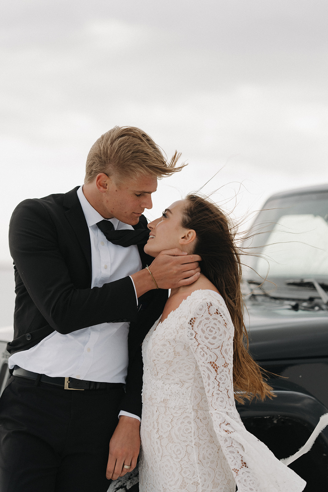 Newlyweds lean against a black jeep while going in for a kiss on a windy day