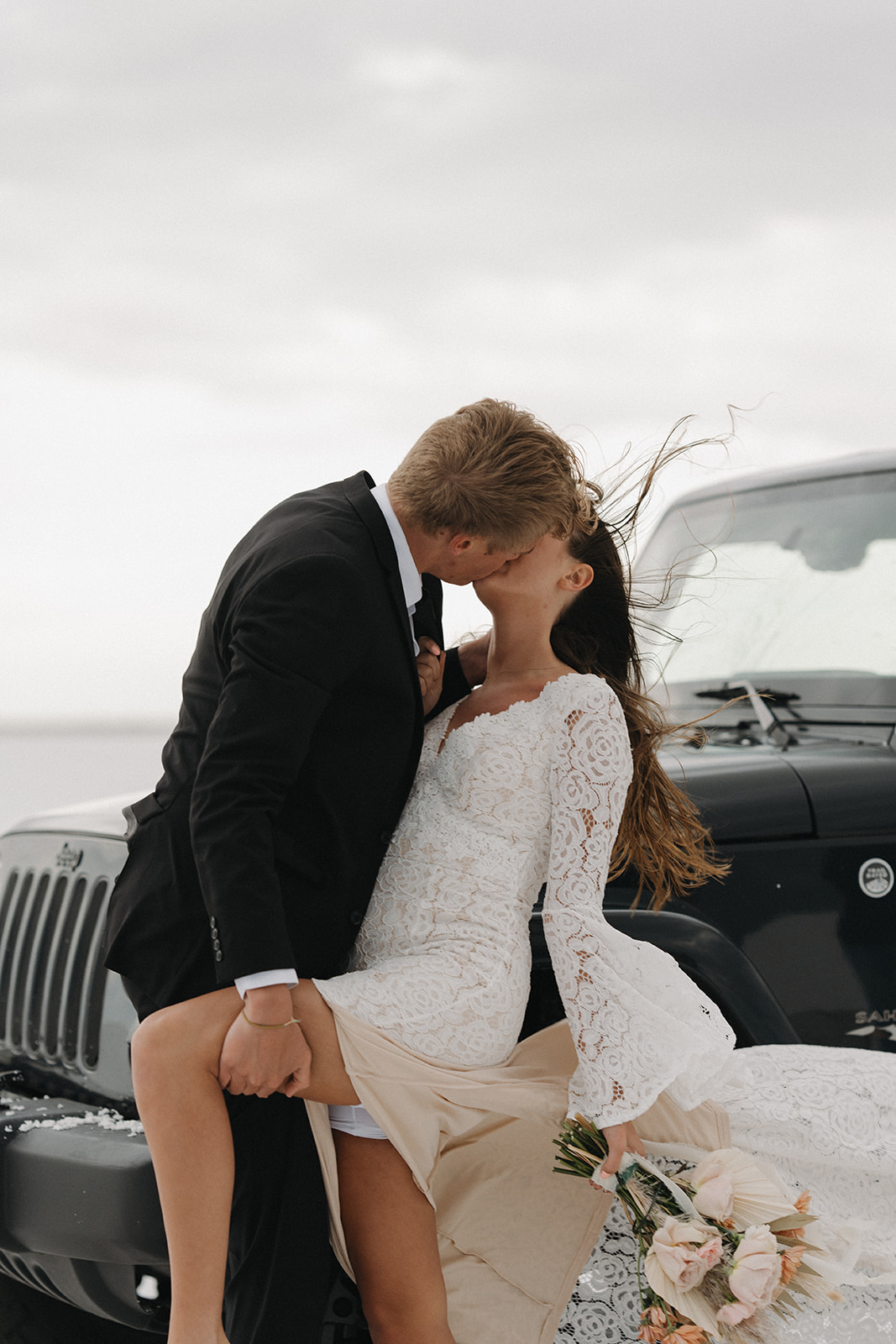 A groom holds the leg of his bride as they kiss leaning against a black jeep during their Salt Flats Elopement