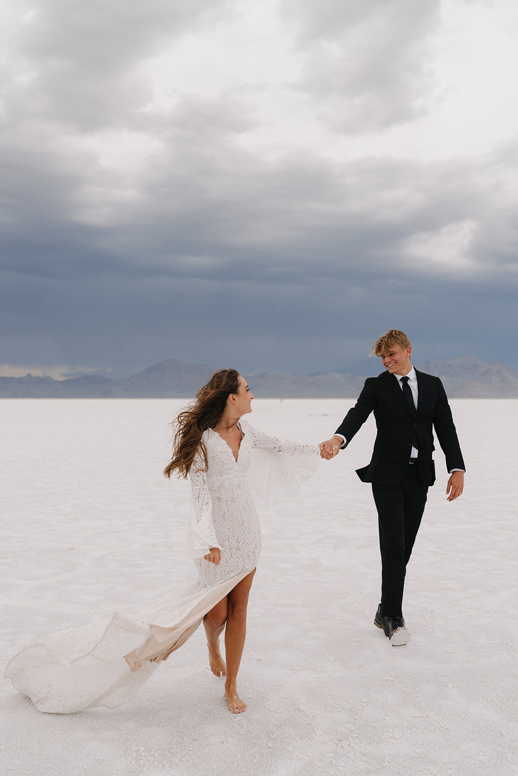 Newlyweds walk hand in hand through the Salt Flats in a black suit and white lace dress flowing in the wind during their Salt Flats Elopement