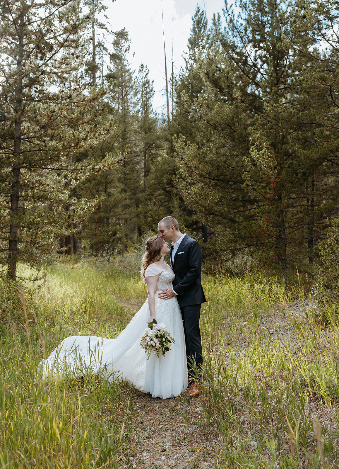 A groom kisses his bride's forehead as they stand in a grassy mountain trail