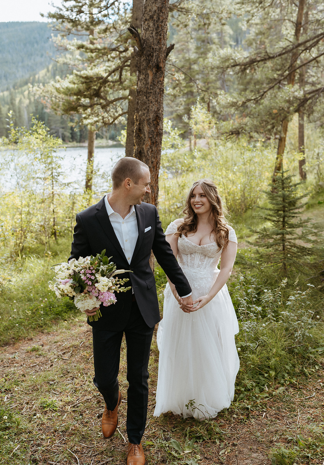 A groom holds the colorful bouquet and leads his wife through a mountain trail on the edge of a lake