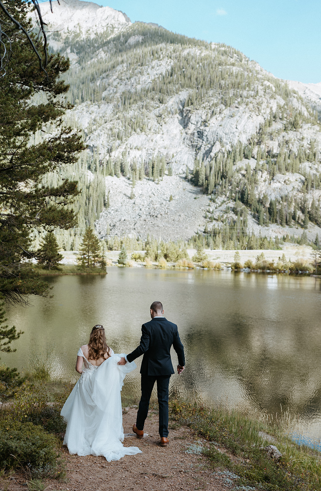 A groom helps his bride down a lakeside mountain trail Colorado Adventure Elopement