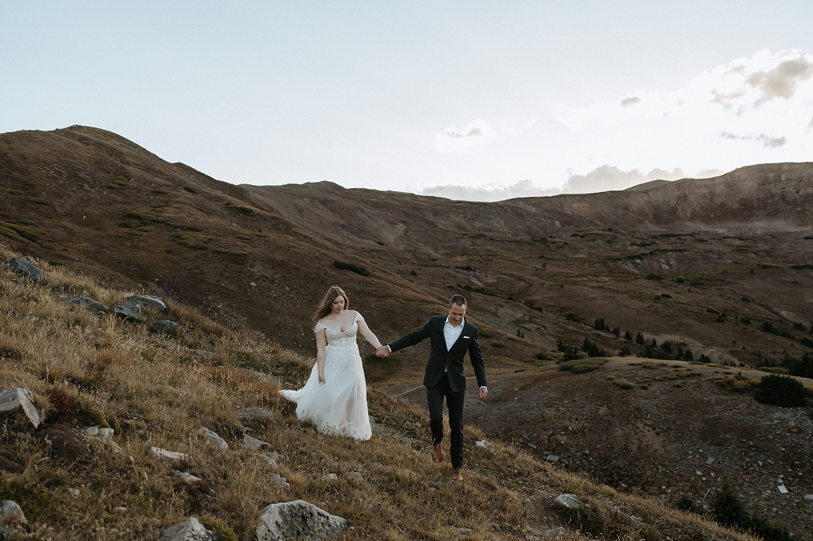 A groom in a black suit walks his bride on a mountain trail leading her by the hand during their Colorado Adventure Elopement