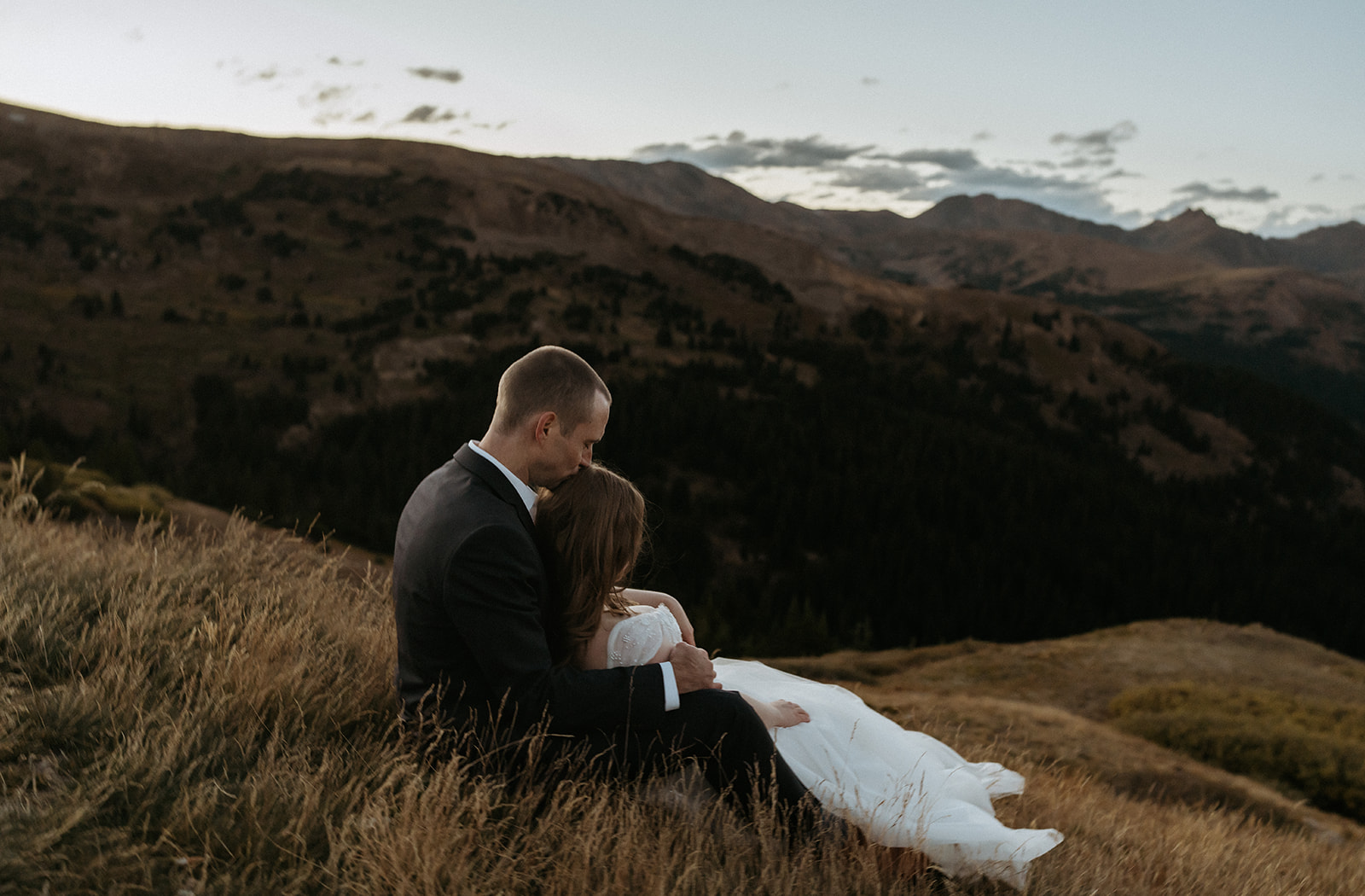 Newlyweds sit on a hillside with the bride in the groom's lap as he kisses her head at sunset during their Colorado Adventure Elopement