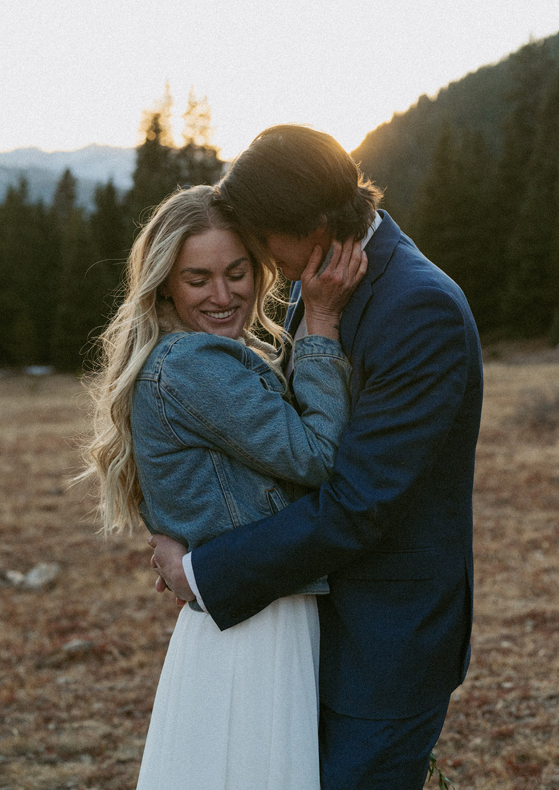 A bride in a white dress and denim jacket is nuzzled by her husband in a remote field
