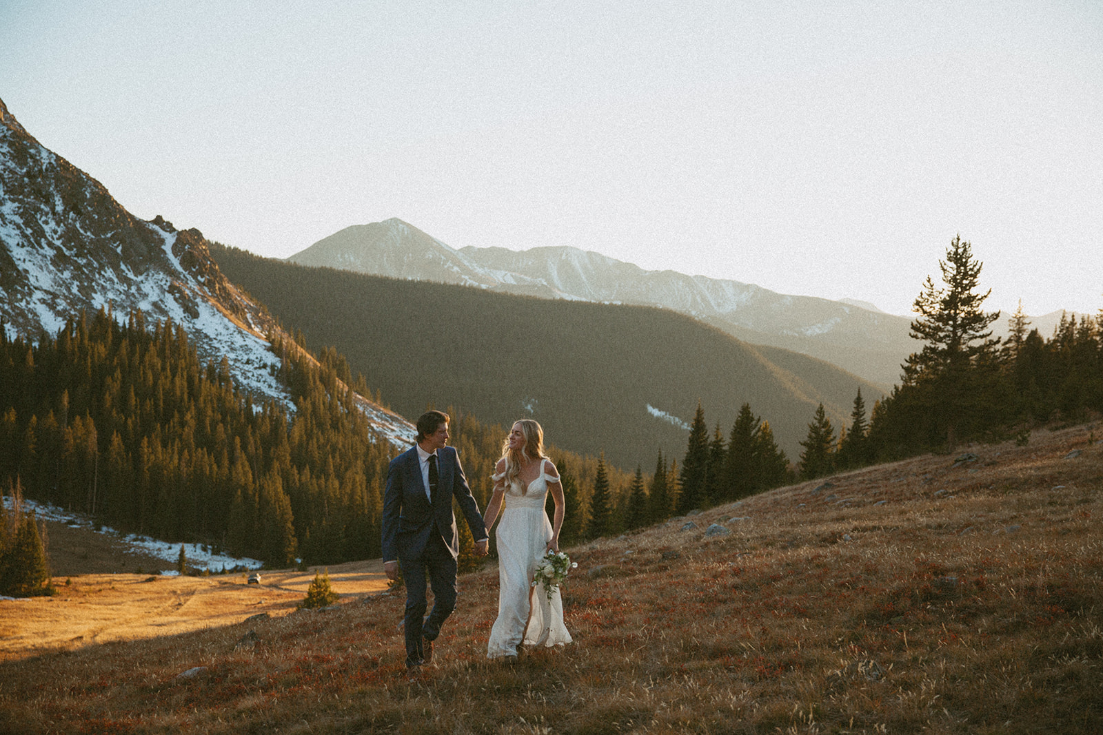 Newlyweds walk hand in hand up a remote hill at sunset during their Colorado Helicopter Wedding