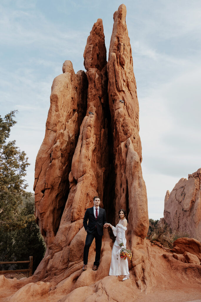 Bride and groom posing for photos in front of a large red rock formation in Garden of The Gods. 