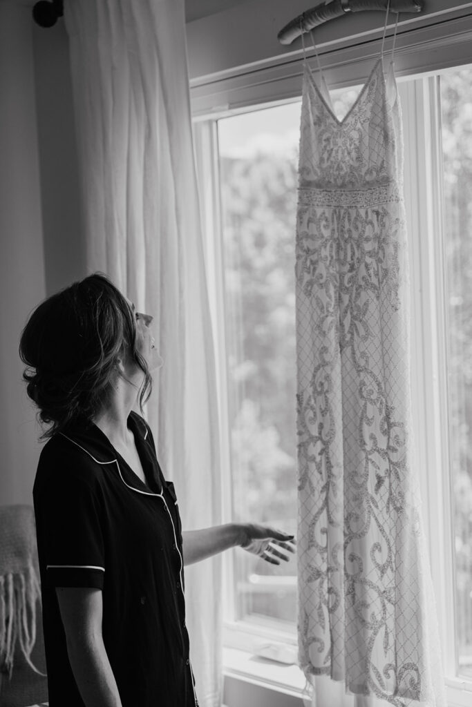 Black and white photo of a bride admiring her hanging wedding dress