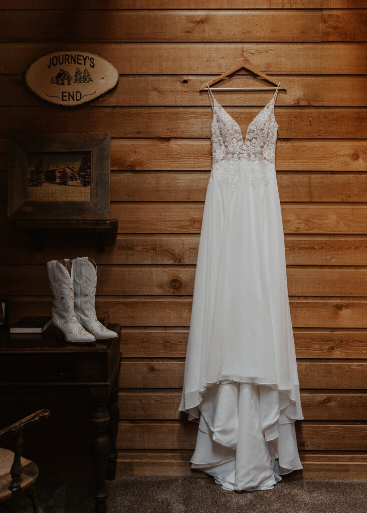 Brides wedding dress hanging next to her white cowgirl boots at a rustic mountain house