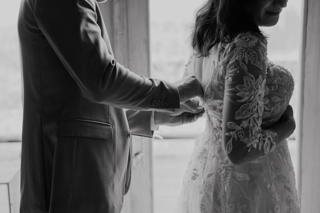 Black and white photo of a groom helping his bride get into her dress