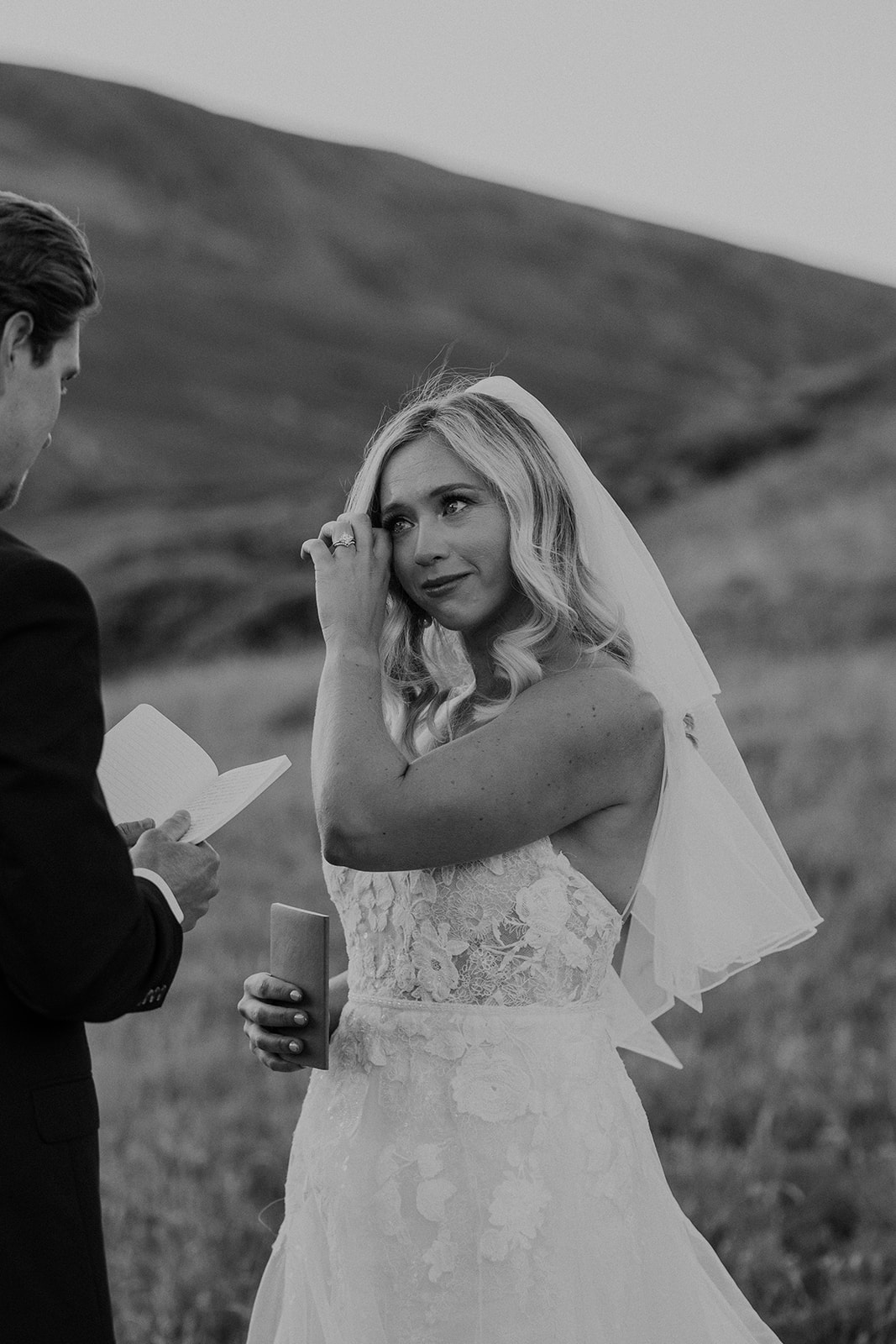 Black and white photo of a bride getting emotional as her groom reads his vows to her